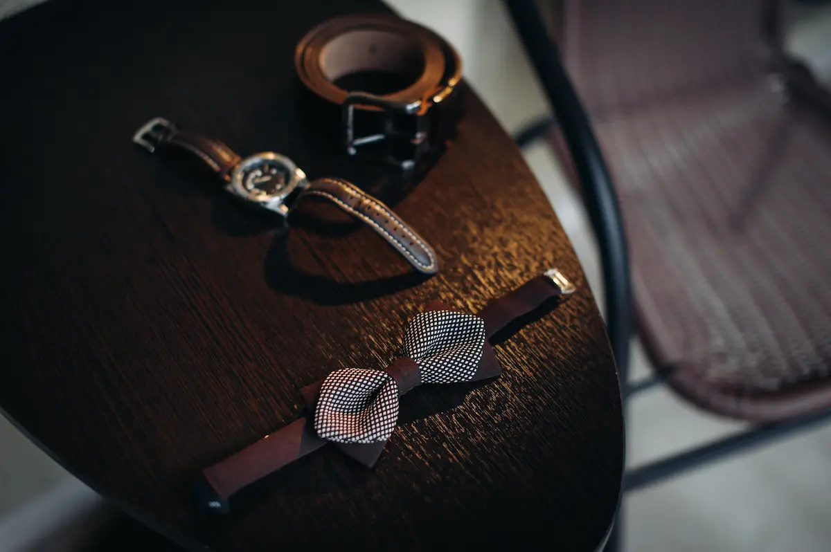 Luxury fashion accessories including a bow tie, leather belt, and wristwatch arranged on a wooden table.